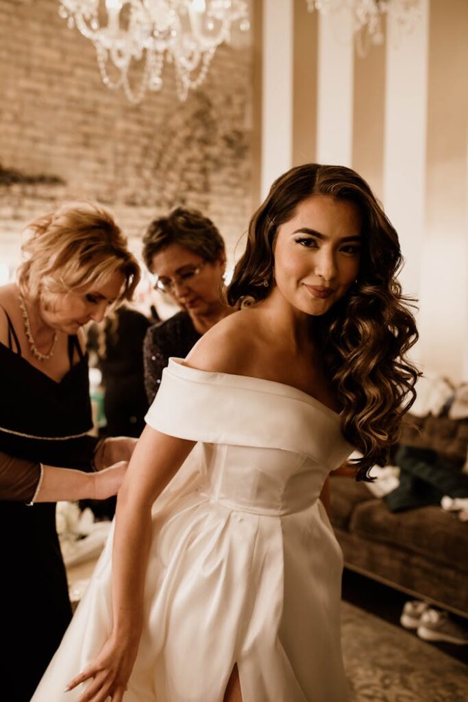 A bride with long wavy hair in a stunning white dress, preparing for her wedding with assistants indoors.