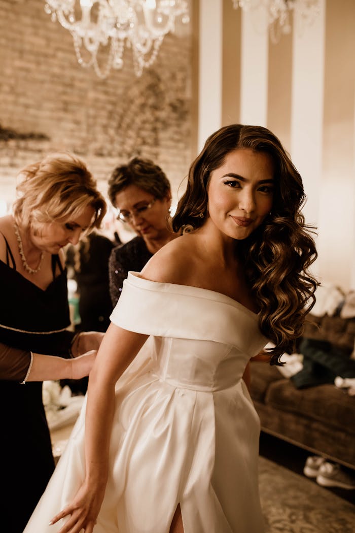 A bride with long wavy hair in a stunning white dress, preparing for her wedding with assistants indoors.