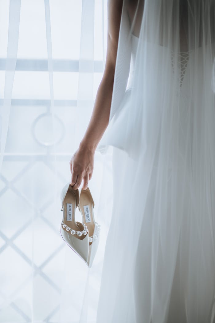 A bride holding elegant bridal shoes adorned with pearls in gentle, soft light.